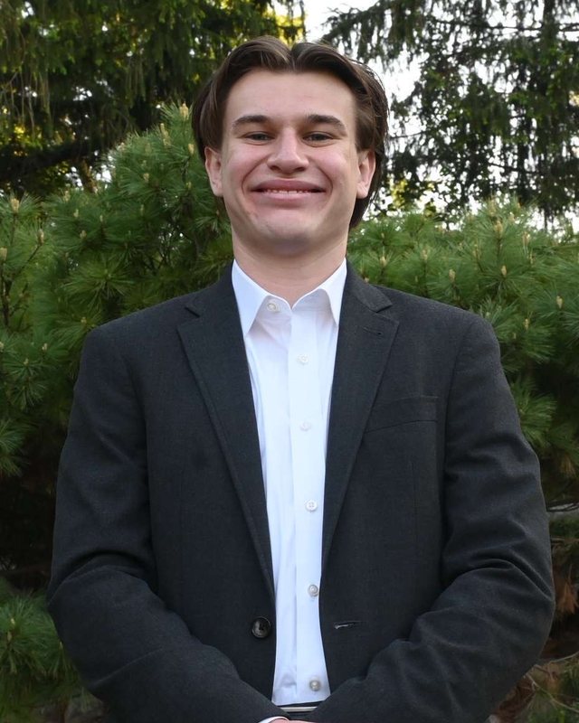 James Hawkins in a dark blazer and white shirt, smiling outdoors in front of pine trees